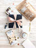 Gift box with pink ribbon, 'One More Cocoa' chocolate bar, and 'Candide' product on a wooden surface.