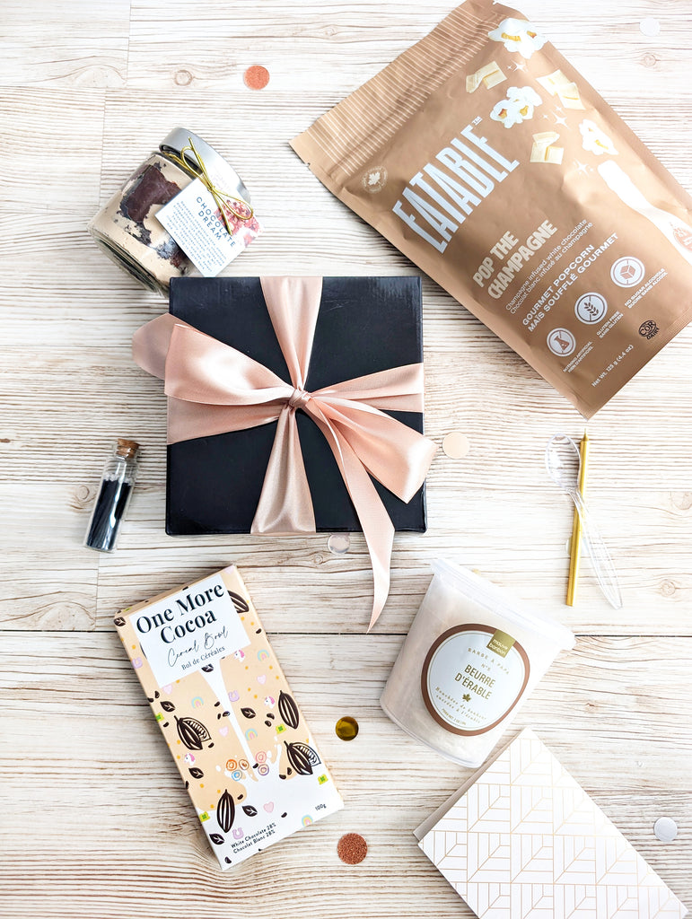 Gift box with pink ribbon, 'One More Cocoa' chocolate bar, and 'Candide' product on a wooden surface.